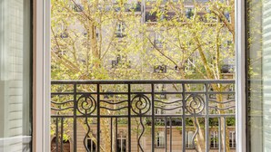Ein Balkon mit schmiedeeisernem Geländer, der auf eine von Bäumen gesäumte Straße und Häuser blickt.