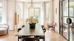 Ein Essbereich mit einem langen Tisch, Stühlen, einer weißen Blumenvase und großen Fenstern mit Vorhängen.