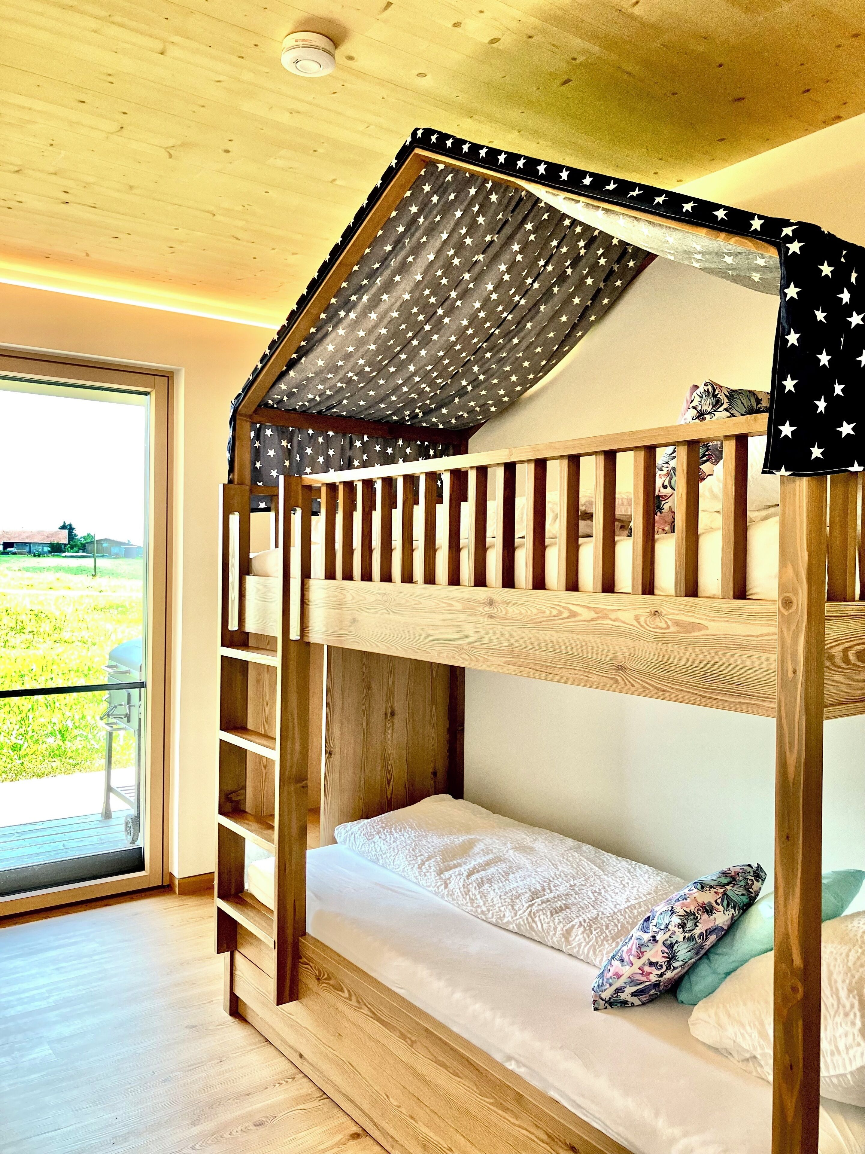 A bunk bed with a star-patterned canopy, wooden headboard, and a view of a grassy area through a large window.