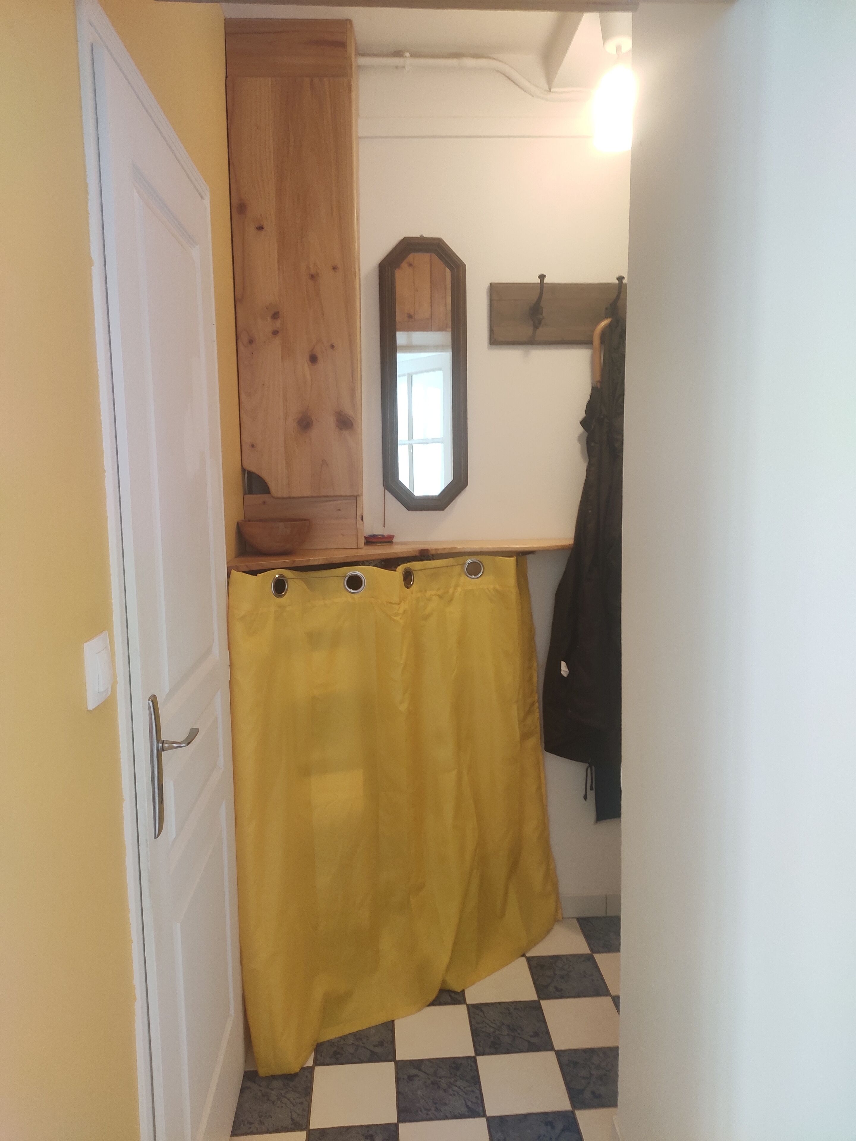 Un estrecho pasillo con piso de cuadros, una puerta de madera, una cortina amarilla, un espejo y un perchero.