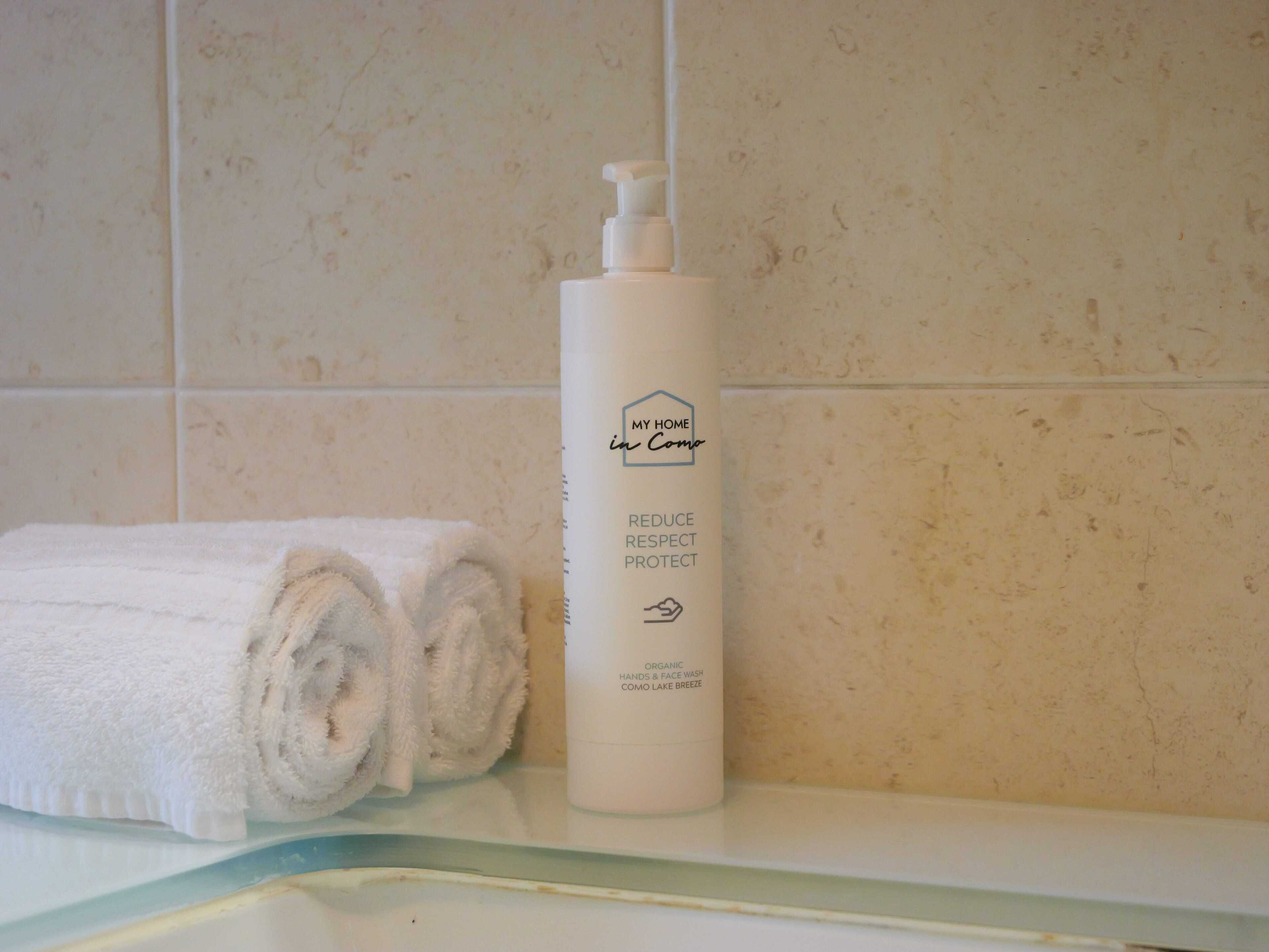 A bathroom with a white towel rolled up on the counter, a white bottle of organic hand and face wash, and a tiled wall.
