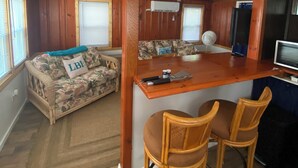Un espace de vie compact comprenant un coin cuisine, un canapé recouvert d’un tissu à motifs floraux et un coin repas en bois avec deux chaises.
