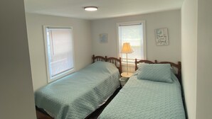 Une chambre avec deux lits simples, des têtes de lit en bois, une petite table de chevet avec une lampe et une fenêtre avec des stores.