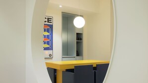 Een modern interieur met een gele tafel en donkerblauwe stoelen, een witte muur met een geometrisch kunstwerk, en een hanglamp.