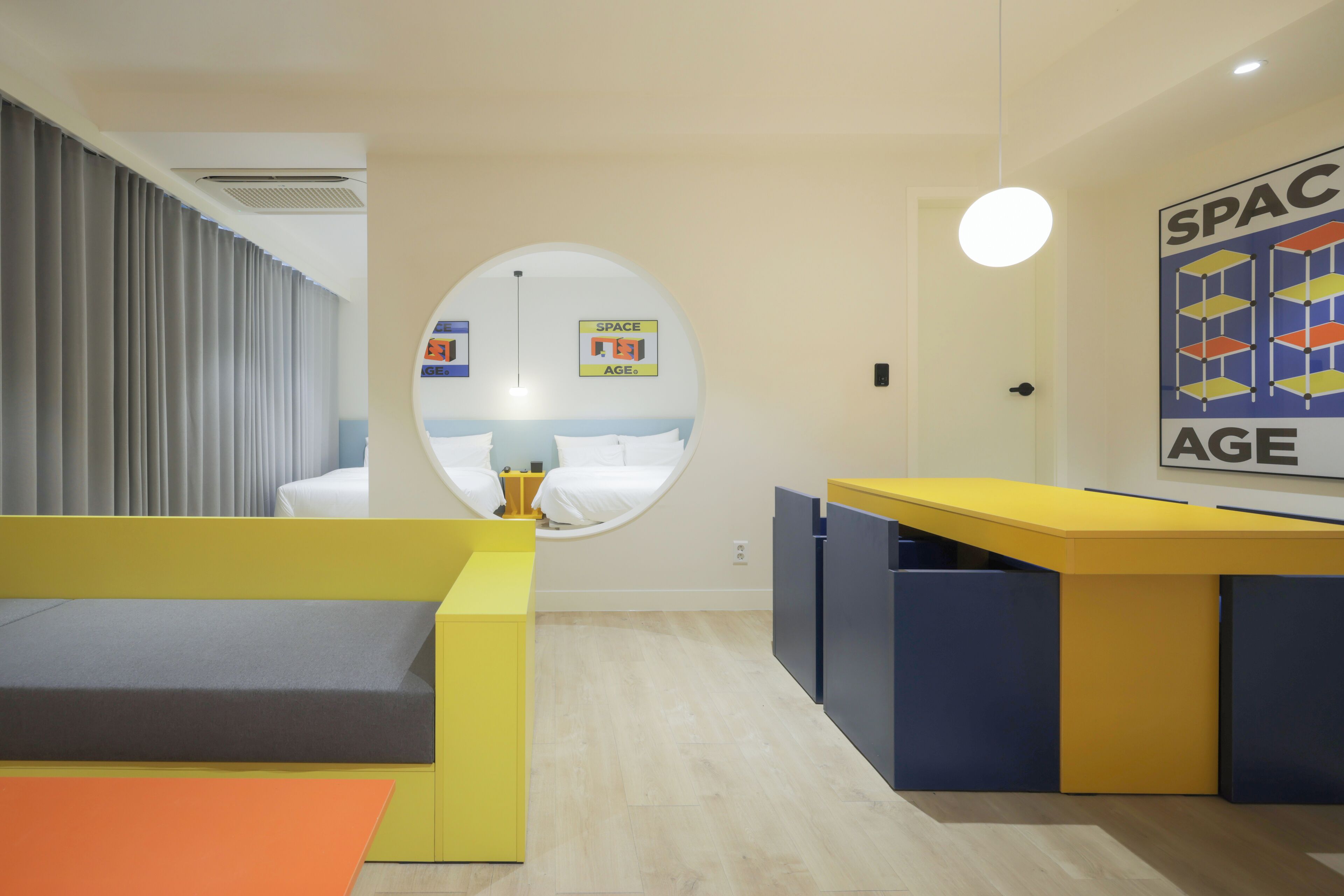 Una habitación moderna con un ventanal circular, un escritorio amarillo y azul, y un sofá gris.