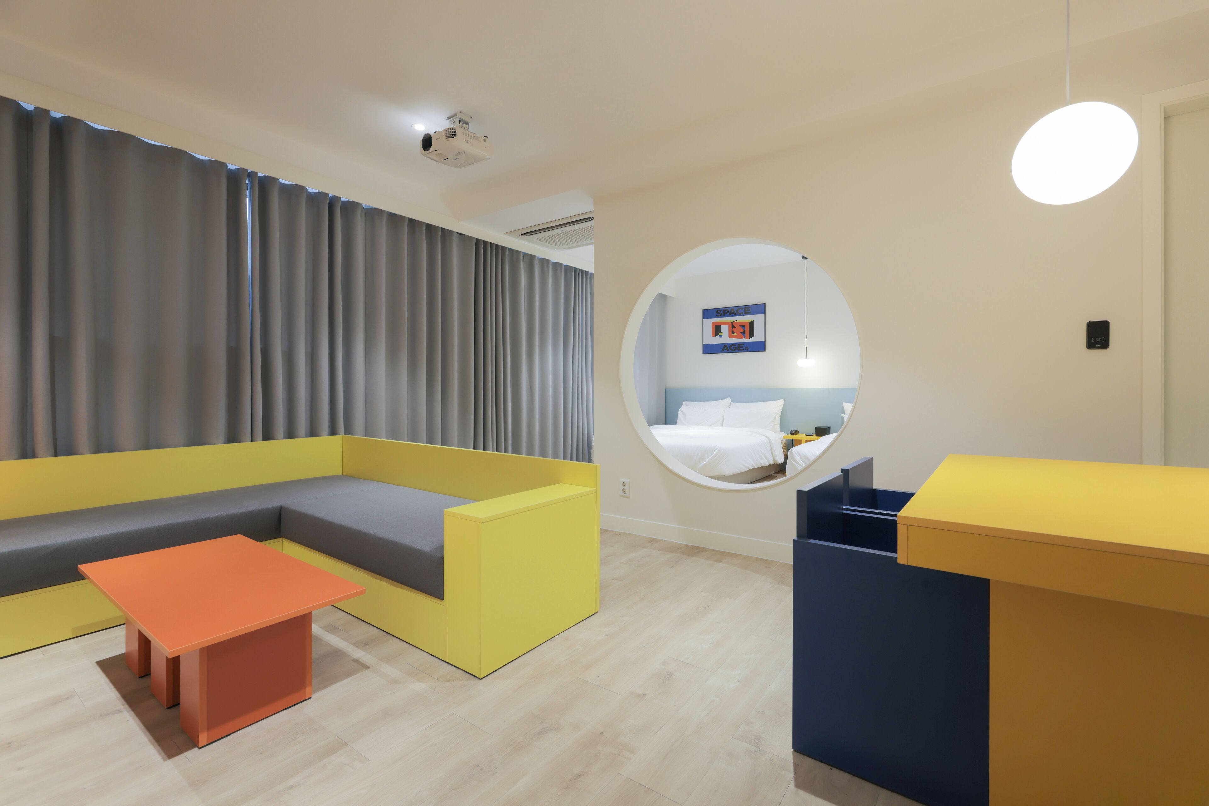 Una habitación de hotel moderna con un sofá amarillo, una mesita naranja pequeña y un espejo circular en la pared.