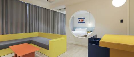Una habitación de hotel moderna con un sofá amarillo, una mesita naranja pequeña y un espejo circular en la pared.