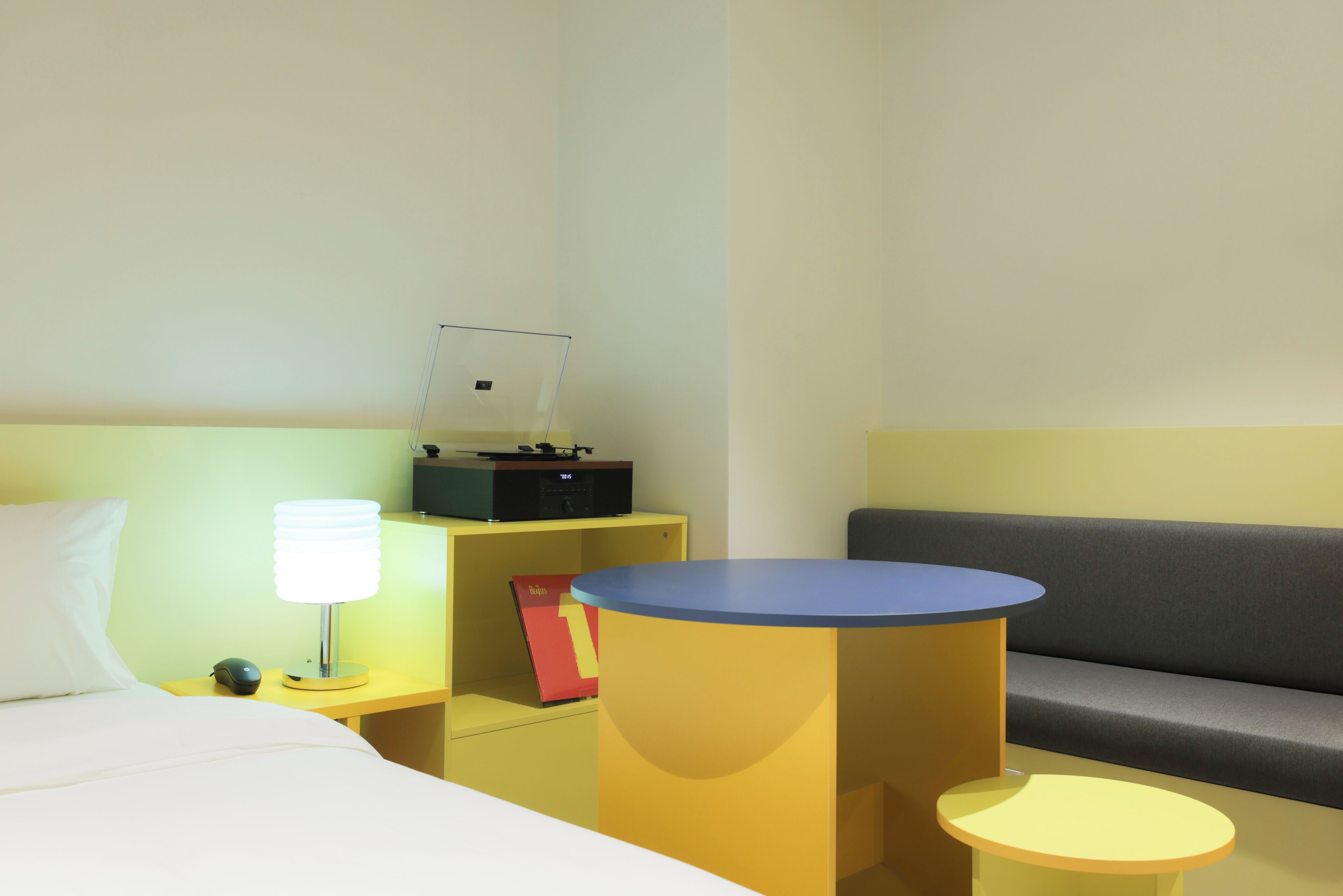 A hotel room with a bed, a desk with a lamp, a small table, a sofa, and a shelf with a book and a box.