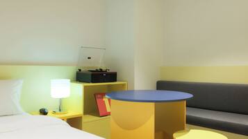 A hotel room with a bed, a desk with a lamp, a small table, a sofa, and a shelf with a book and a box.