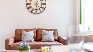 Ein braunes Ledersofa mit zwei gemusterten Kissen, einer Wanduhr und einem Couchtisch mit zwei Gläsern Orangensaft.