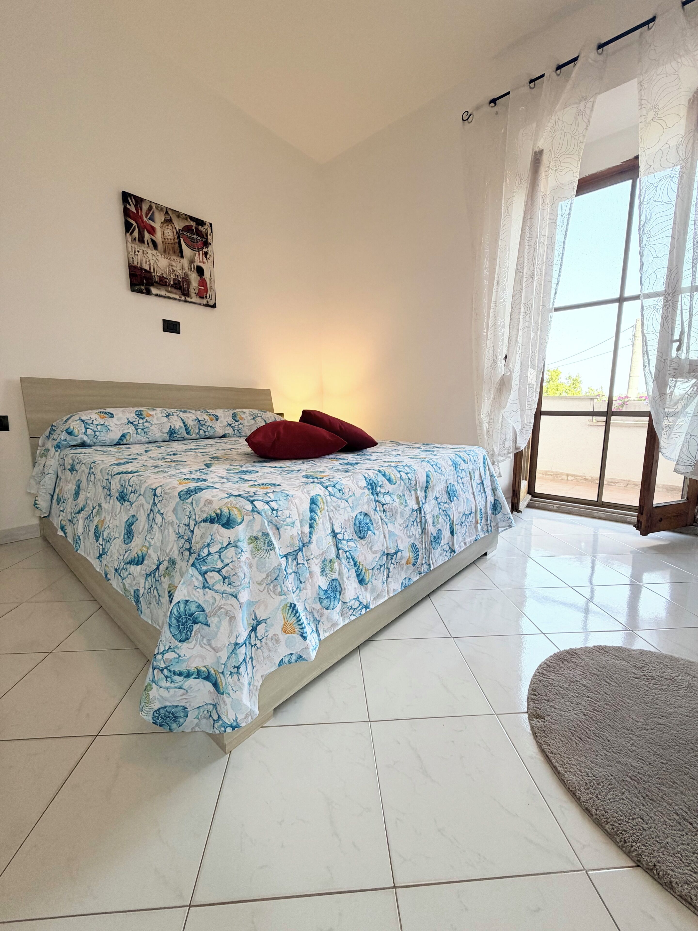 Une chambre à coucher avec un lit, une fenêtre avec des rideaux, un tableau accroché au mur et un sol carrelé.
