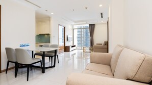 A modern living room with a beige sofa, a dining table with chairs, a kitchen area, and a large window with a view of buildings. - Vinhomes Central Park Luxury - Leminbo (Ho Chi Minh City)