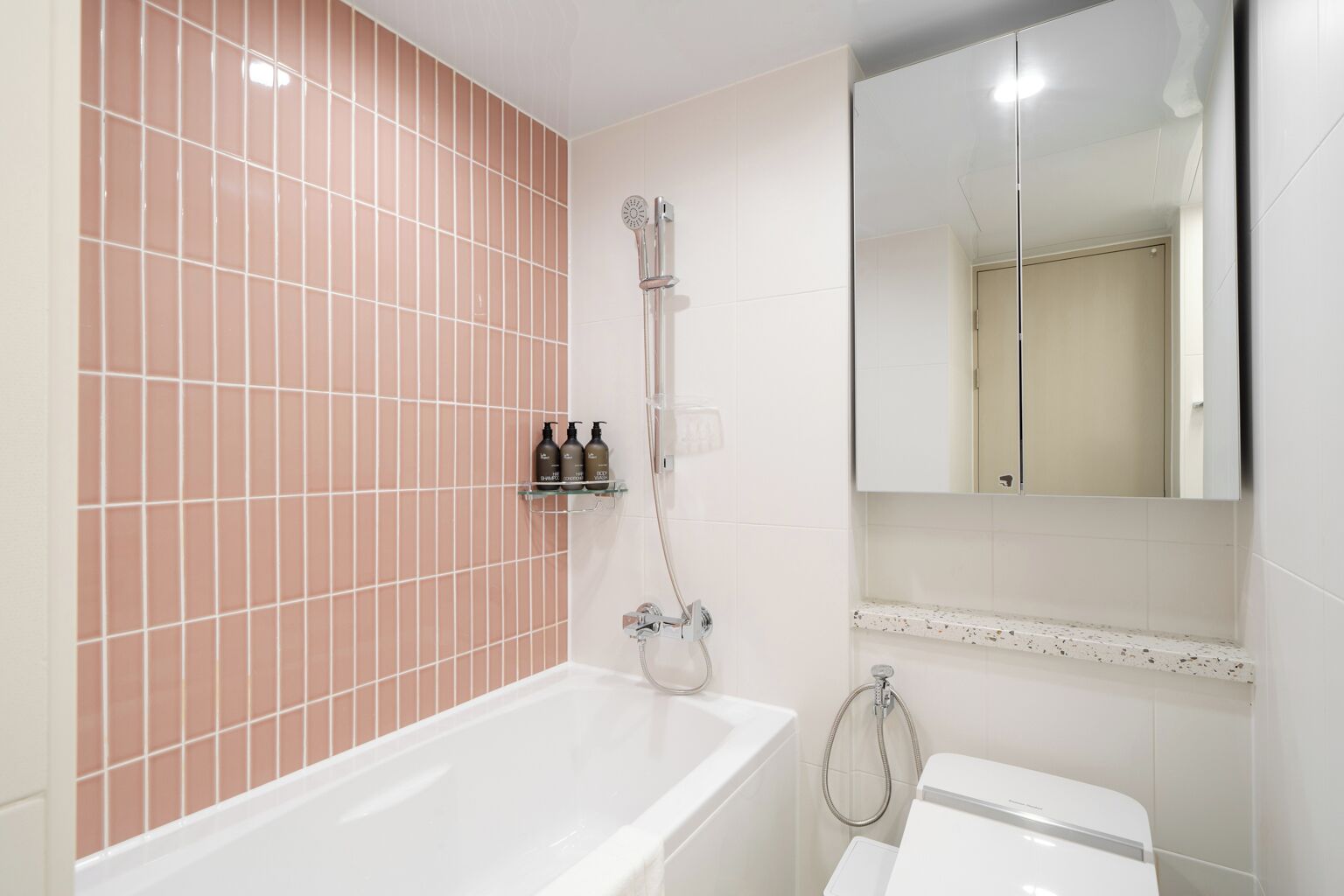 A bathroom with a white bathtub, a shower with a handheld showerhead, and a toilet. There is a shelf with toiletries and a mirror above the sink.