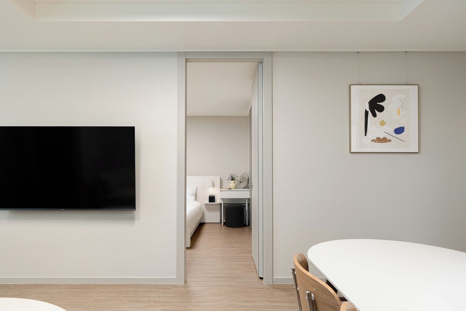 A modern hotel room with a large flat-screen TV, a bed, a desk with a chair, and a small kitchenette.