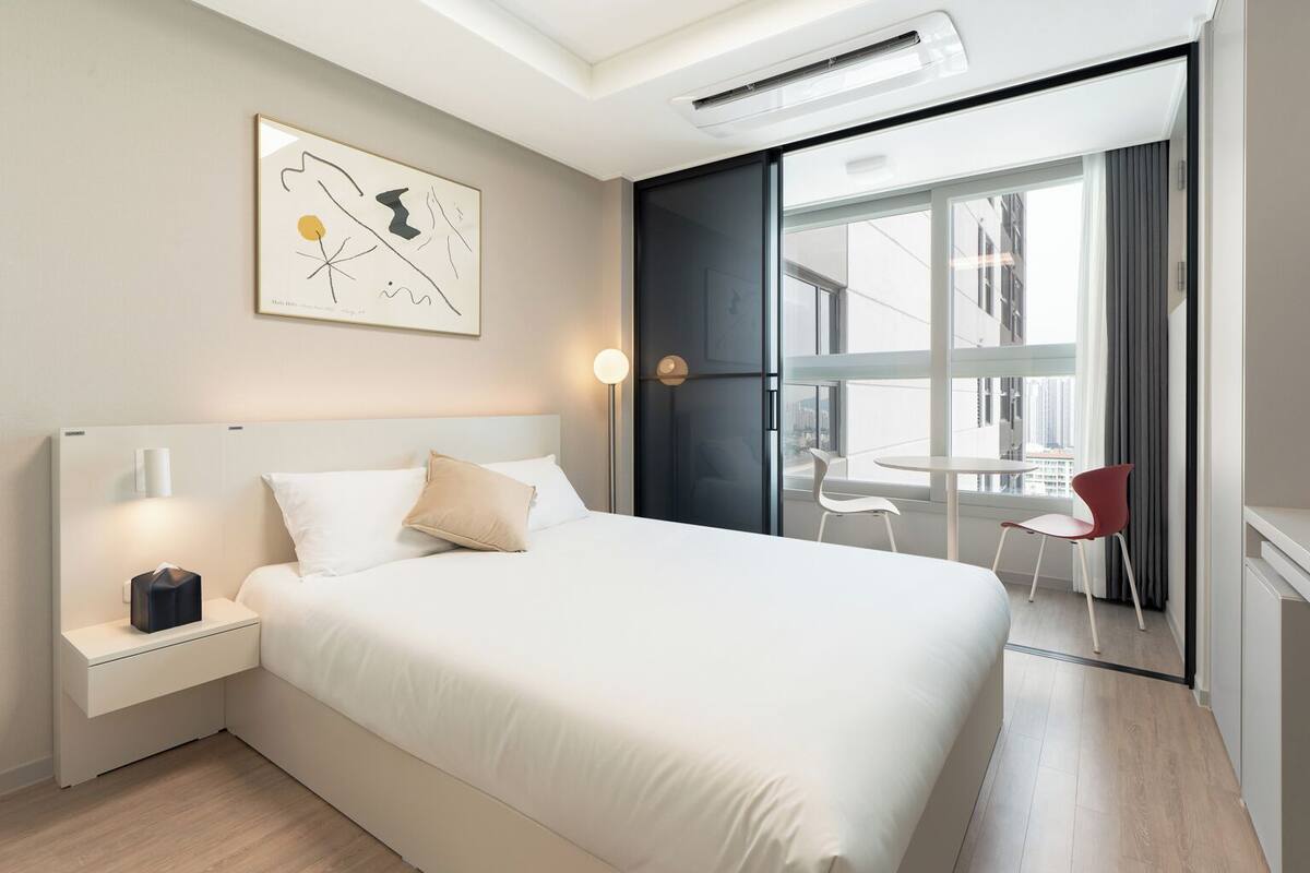 A modern hotel room with a large bed, bedside tables, a wall-mounted artwork, a window with a view of the city, and a balcony with chairs.