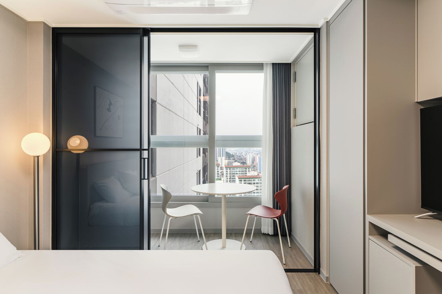 Habitación de hotel moderna con cama, una mesita redonda, dos sillas, televisor y ventana grande con vistas a la ciudad.