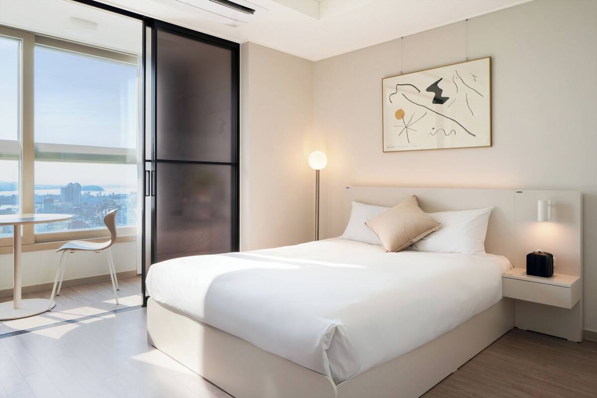 A modern hotel room with a large bed, a desk with a chair, a view of the city through large windows, and a minimalist artwork on the wall.