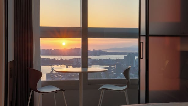 Une pièce dotée d’une grande fenêtre donnant sur un paysage urbain au coucher du soleil, et comprenant deux fauteuils et une petite table.