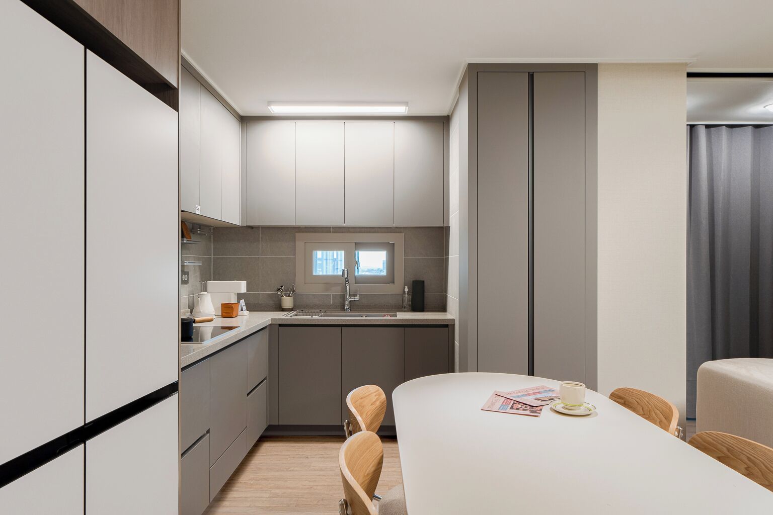 A modern kitchen with grey cabinets, a white countertop, and a window above the sink. There is a dining area with a white table and chairs.