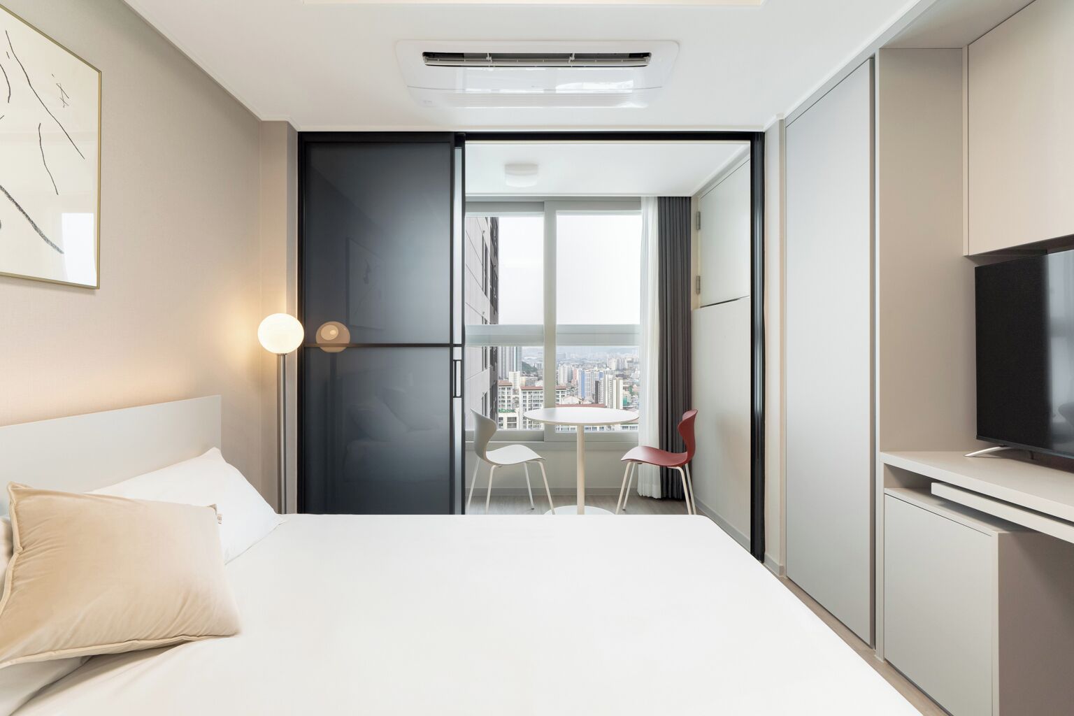 A modern hotel room with a bed, a TV, a balcony with a view of the city, and a dining area with chairs.