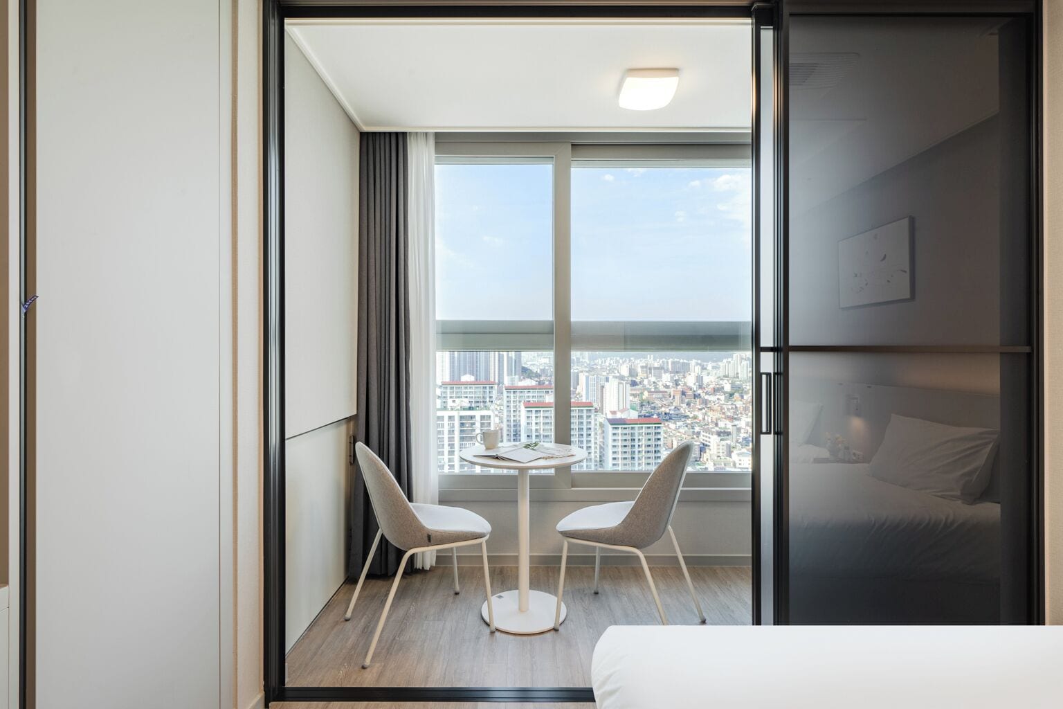 Habitació d'hotel moderna amb un gran finestral que ofereix vista a la ciutat, una taula rodona petita amb dues cadires i un llit amb capçalera.