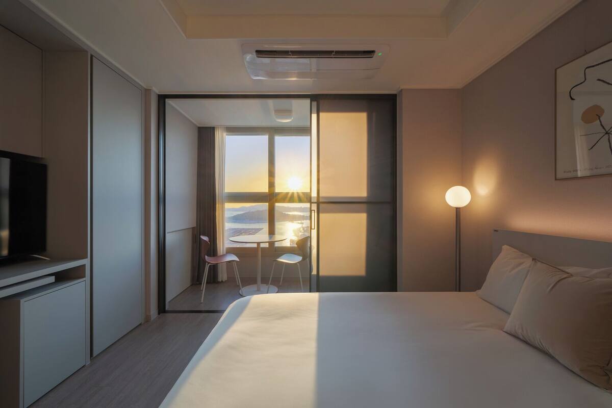 A hotel room with a bed, a TV, a desk with chairs, and a view of the sunset through large windows.