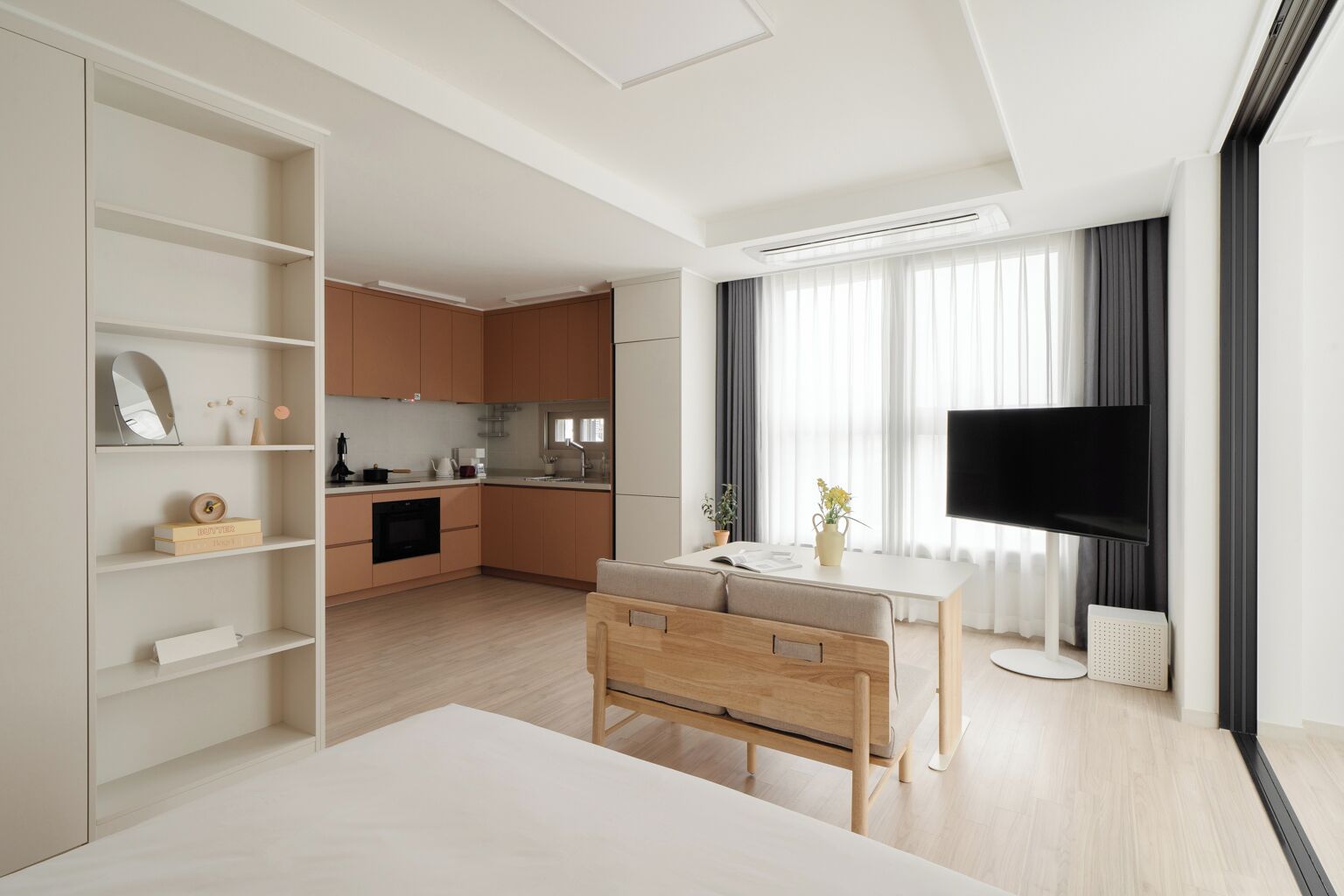 A modern hotel room with a large bed, a desk, a TV, a kitchenette, and a large window with curtains.