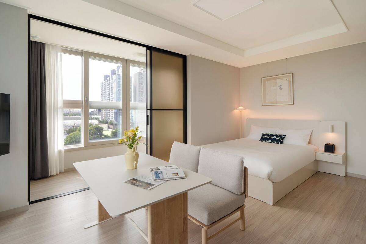 A modern hotel room with a bed, a desk, and a chair. There is a window with curtains, a vase with flowers, and a lamp on the bedside table.