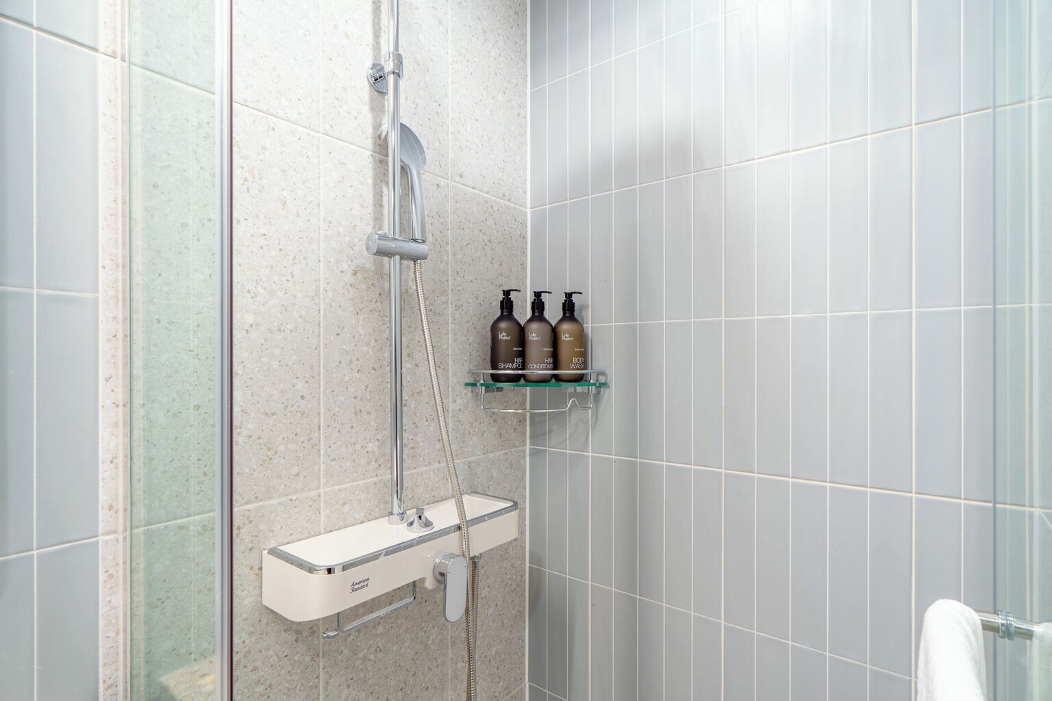A shower area with a handheld showerhead, a wall-mounted faucet, and a glass shelf with three bottles of shampoo and conditioner.