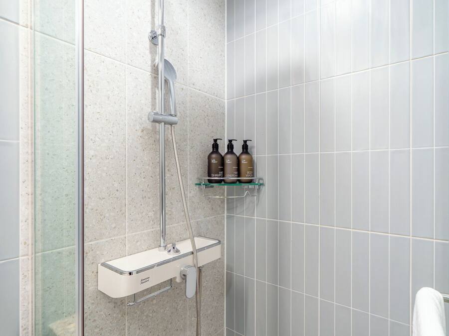A shower area with a handheld showerhead, a wall-mounted faucet, and a glass shelf with three bottles of shampoo and conditioner.