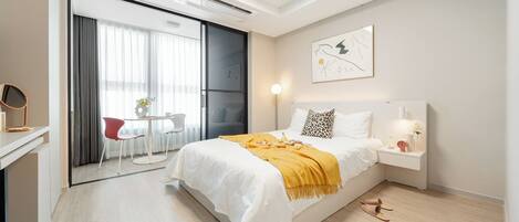 Una habitación de hotel moderna con una cama, mesita de noche y un escritorio con una silla. Hay una puerta corredera que da a un balcón con mesa y sillas.