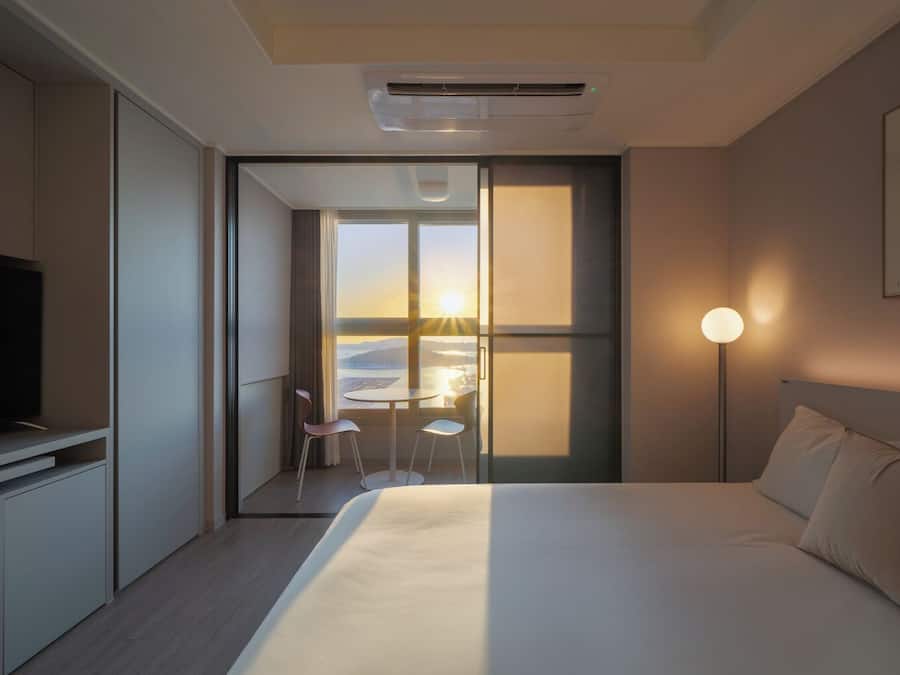 A hotel room with a bed, a TV, a desk with chairs, a lamp, and a view of the sunset through a window.