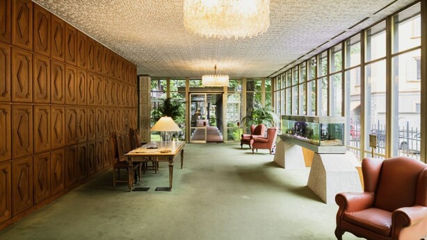 Une pièce spacieuse avec un plafond haut, un lustre, un mur lambrissé, un bureau avec une lampe, une chaise et un aquarium.