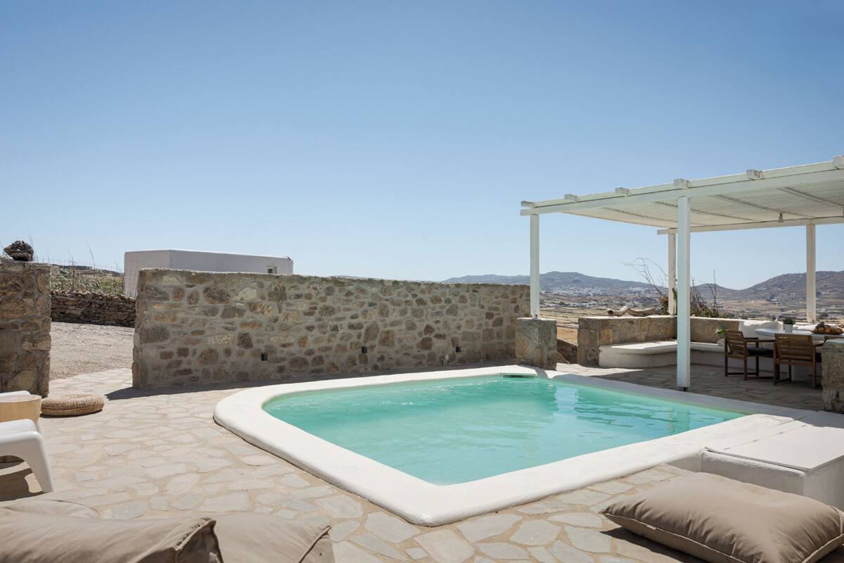 Ein Becken aus Stein mit weißem Rand, umgeben von Steinplatten und beigefarbenen Kissen. Im Hintergrund ist eine weiße Pergola mit Tisch und Stühlen zu sehen.