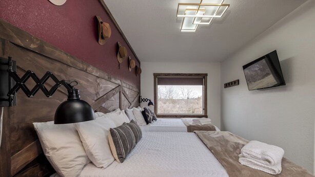 A bedroom with a wooden headboard, multiple pillows, a large bed, a bedside table, a lamp, a window with a view, and a mounted picture. - Double the Comfort | Travel Together (Prescott)