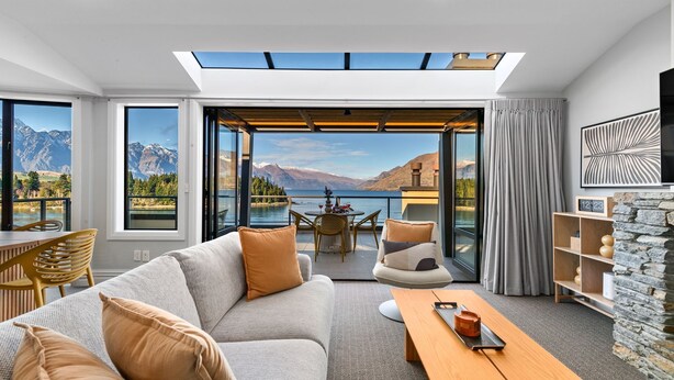 Un salon moderne avec un canapé, une table basse et des fauteuils, donnant sur un lac et des montagnes.