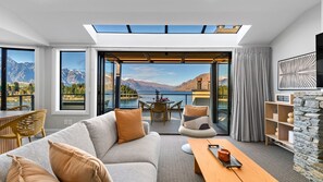 Un salon moderne avec un canapé, une table basse et des fauteuils, donnant sur un lac et des montagnes.