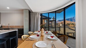 Un espace de repas moderne avec une grande table dressée pour quatre personnes, une cuisine en arrière-plan et une vue sur les montagnes par de grandes fenêtres.