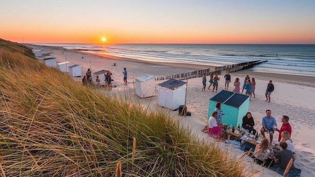 Escena de playa con gente reunida alrededor de una mesa, con vista al océano al atardecer.