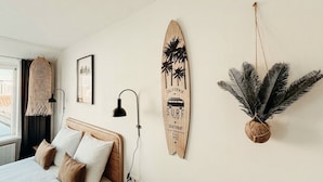 Un dormitorio con una cama, una tabla de surf colgada en la pared, un cuadro enmarco, una planta colgante y una lámpara.