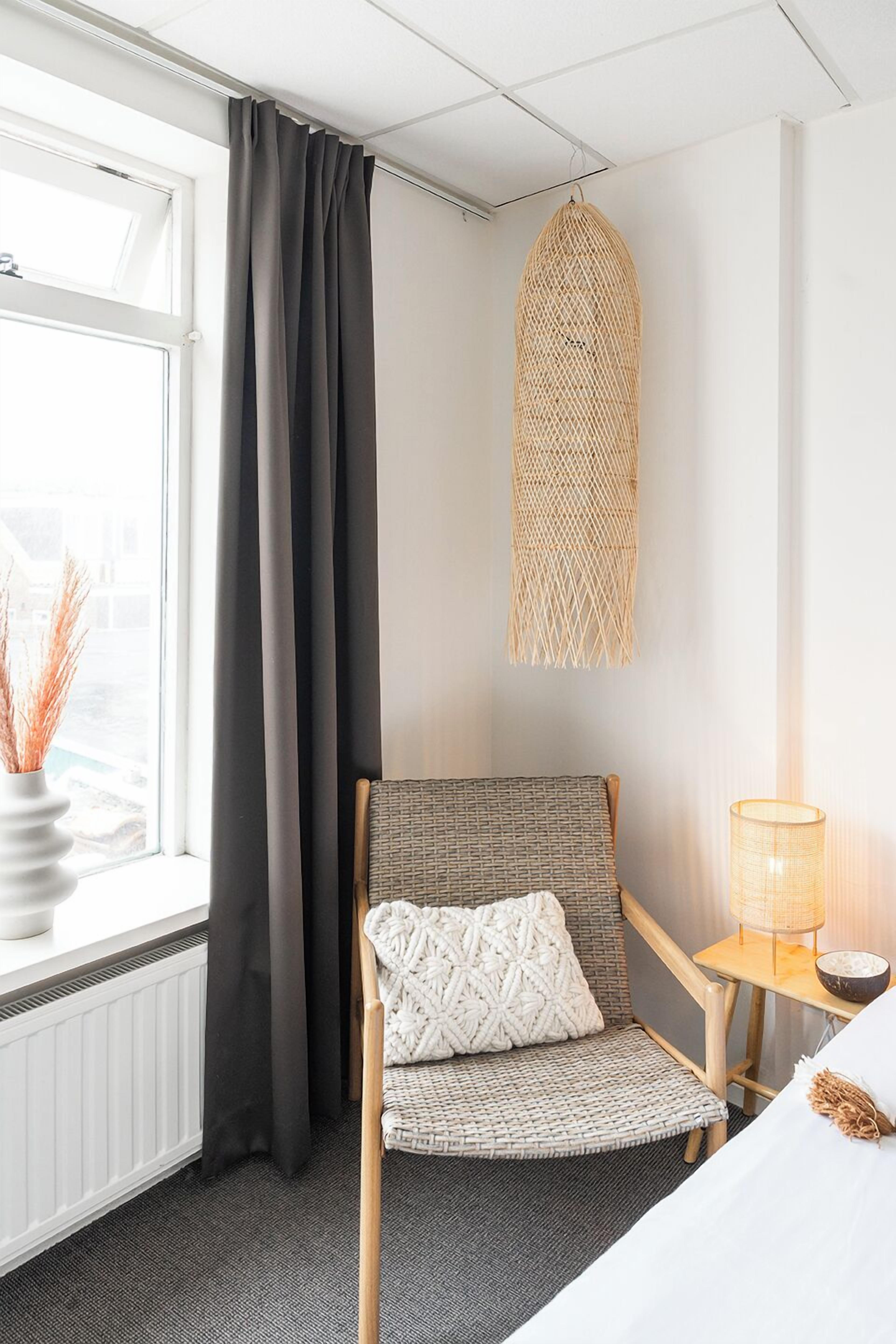 Habitación de hotel con cama, una silla con cojín, una mesita con lámpara, ventana con cortinas y un adorno en la pared.
