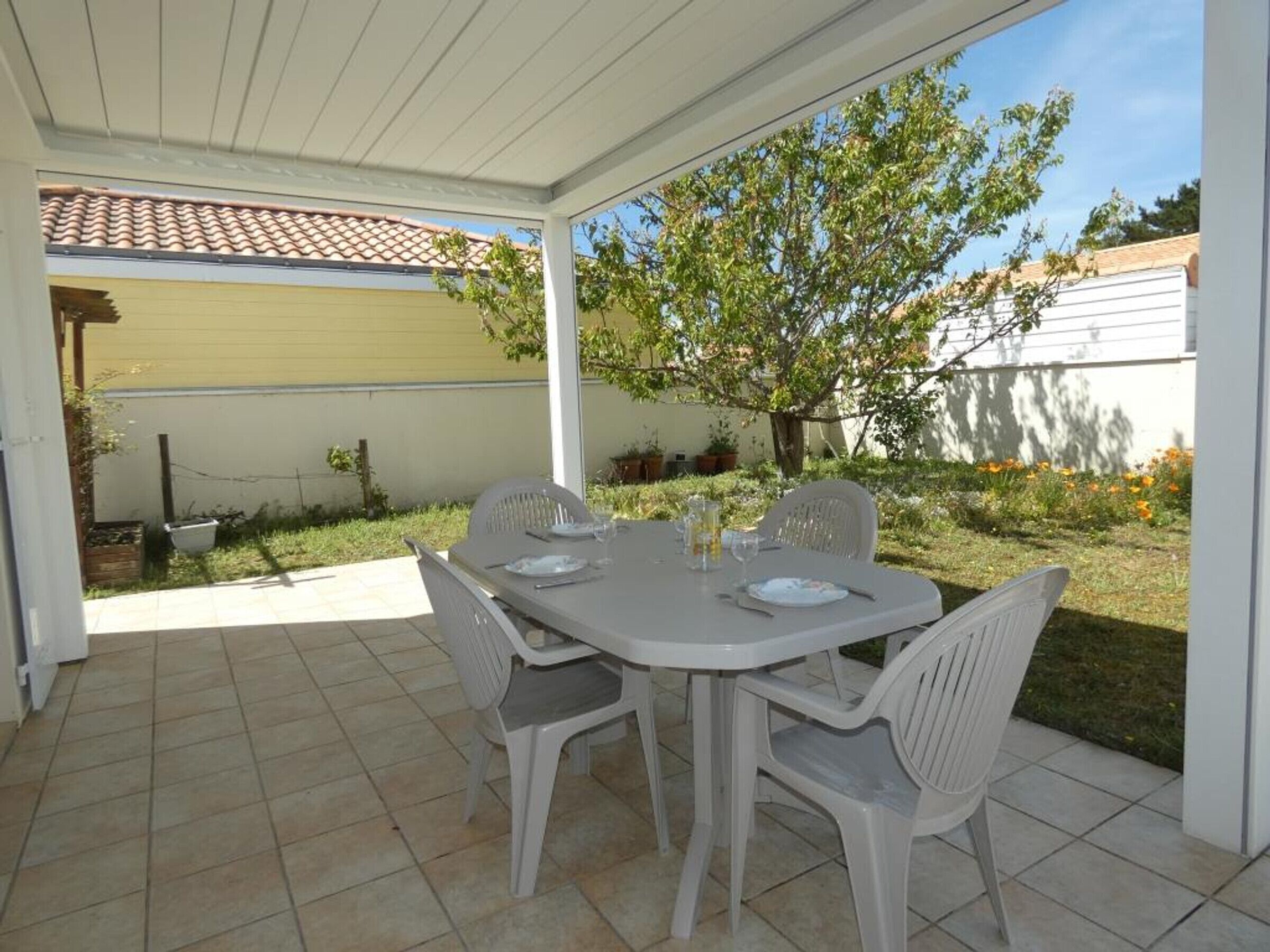 Eine überdachte Terrasse mit Tisch und Stühlen, ein Fliesenboden und Blick auf einen Garten mit Baum und einem Gebäude im Hintergrund.
