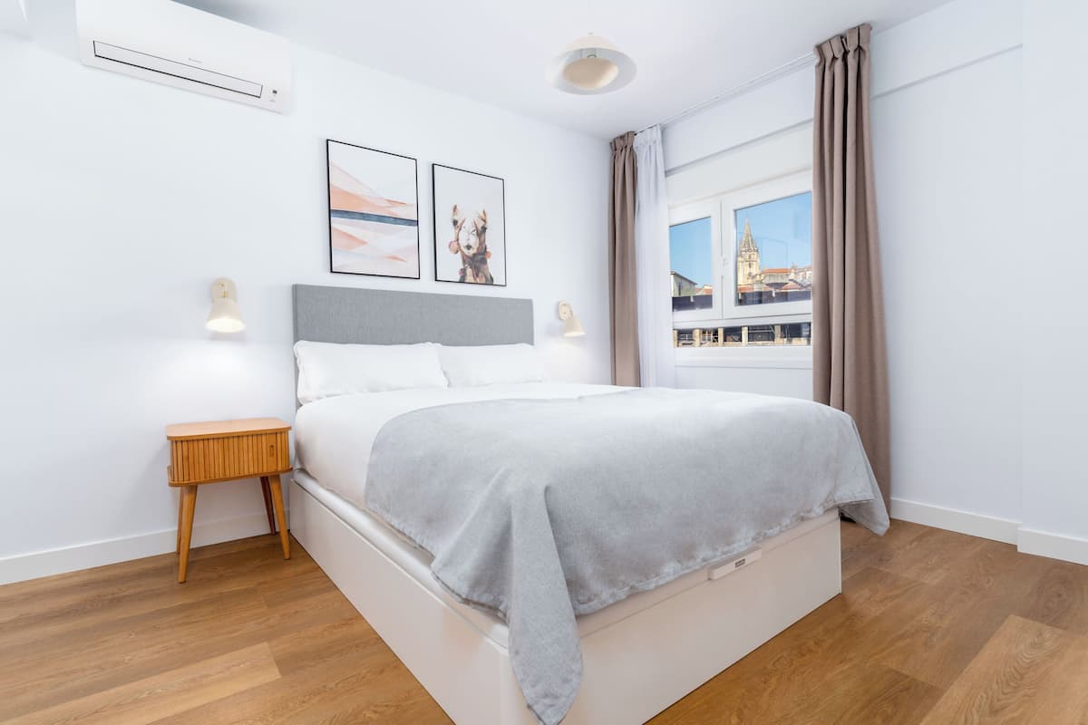 Une chambre moderne avec un grand lit, une table de chevet, une commode et une fenêtre donnant sur un immeuble.
