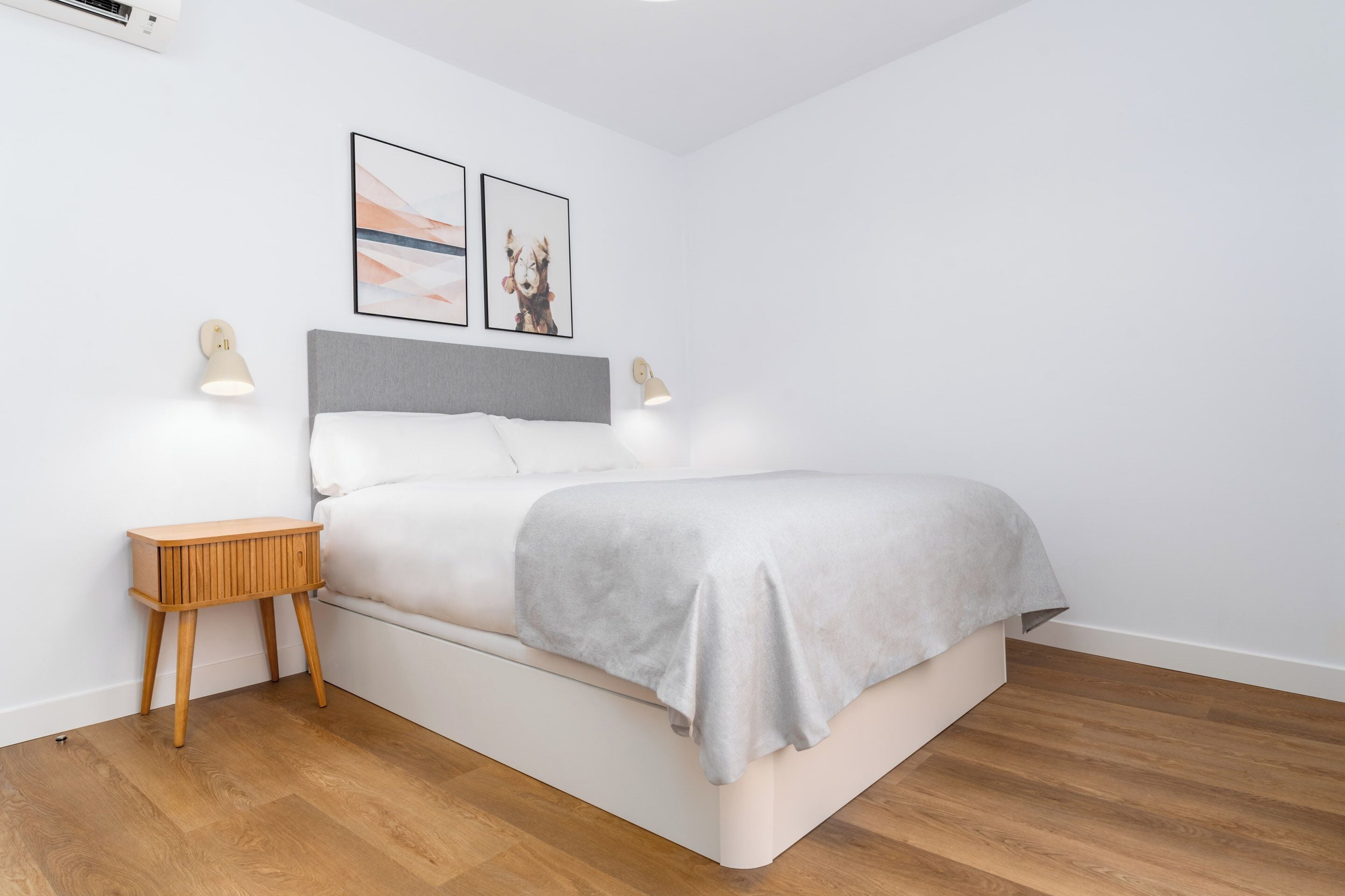 Un dormitorio moderno con cama, mesitas de noche de madera, lámparas de pared y dos cuadros enmarcados.