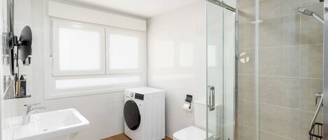 A modern bathroom with a glass shower enclosure, a white sink, a washing machine, and a toilet.