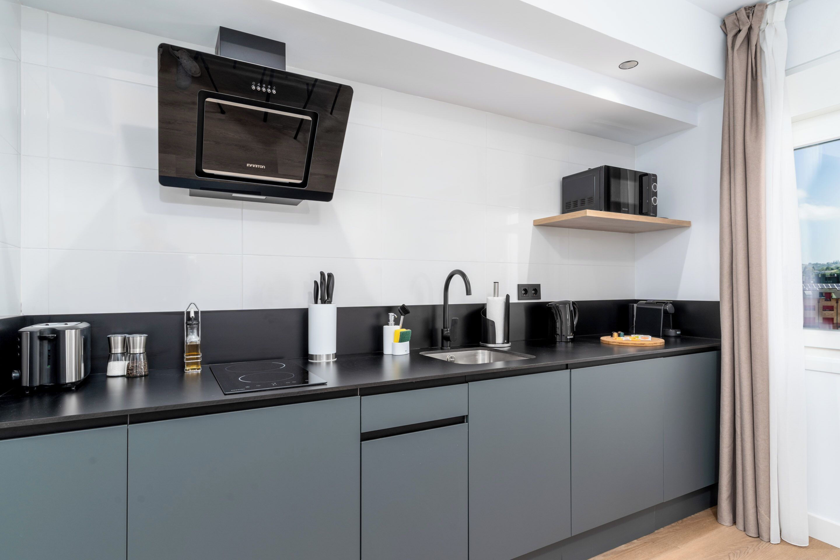 A modern kitchen with grey cabinets, a black countertop, a built-in oven, a microwave, and a window with curtains.