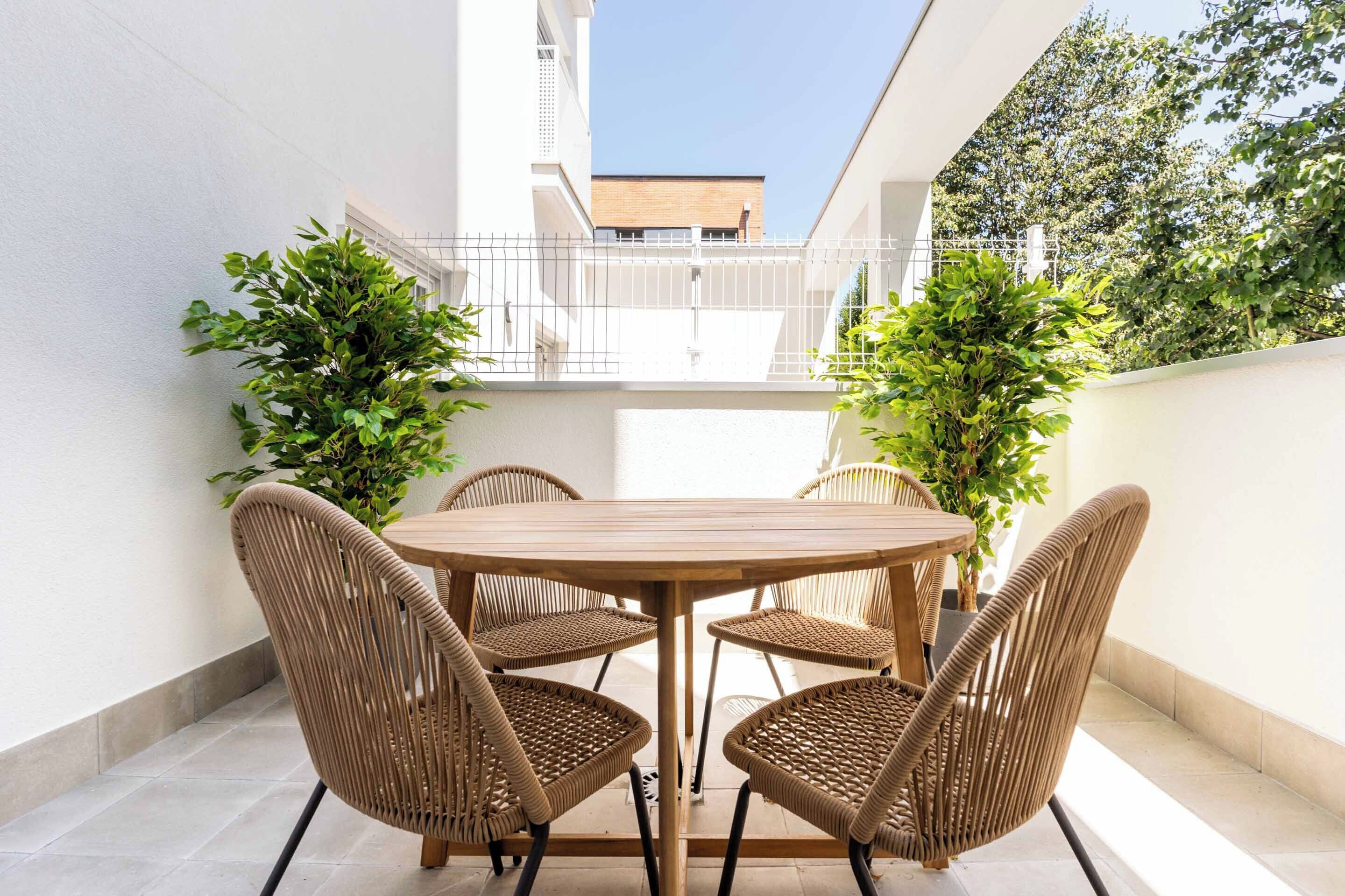 Una mesa redonda de madera con cuatro sillas de mimbre en un espacio exterior iluminado por el sol, con paredes blancas y plantas en macetas.