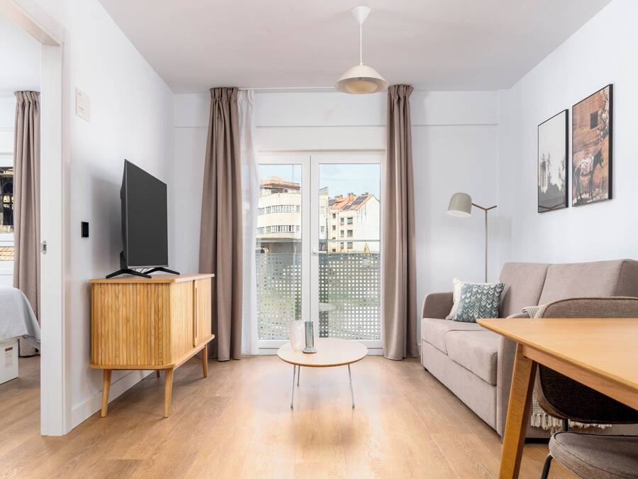 Una sala de estar moderna con suelo de madera, un televisor en un mueble, una mesa redonda, un sofá y vistas a edificios a través de grandes ventanales.