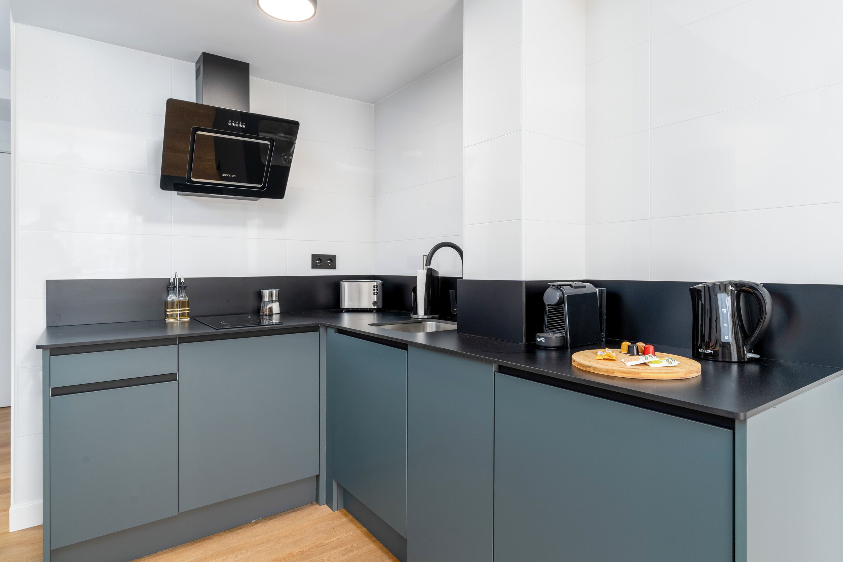 A modern kitchen with grey cabinets, a black countertop, a microwave, a kettle, and a cutting board with food items.