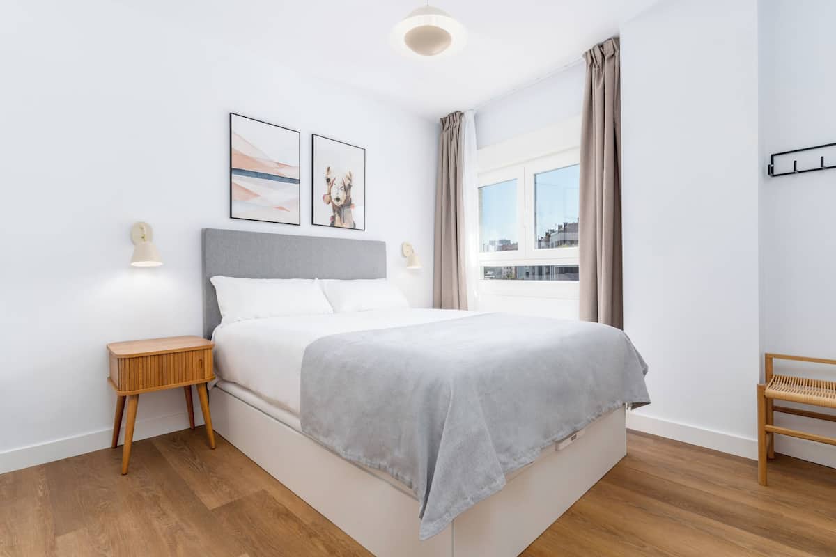 Une chambre à coucher moderne avec un lit, une table de chevet, une lampe et une fenêtre avec des rideaux.